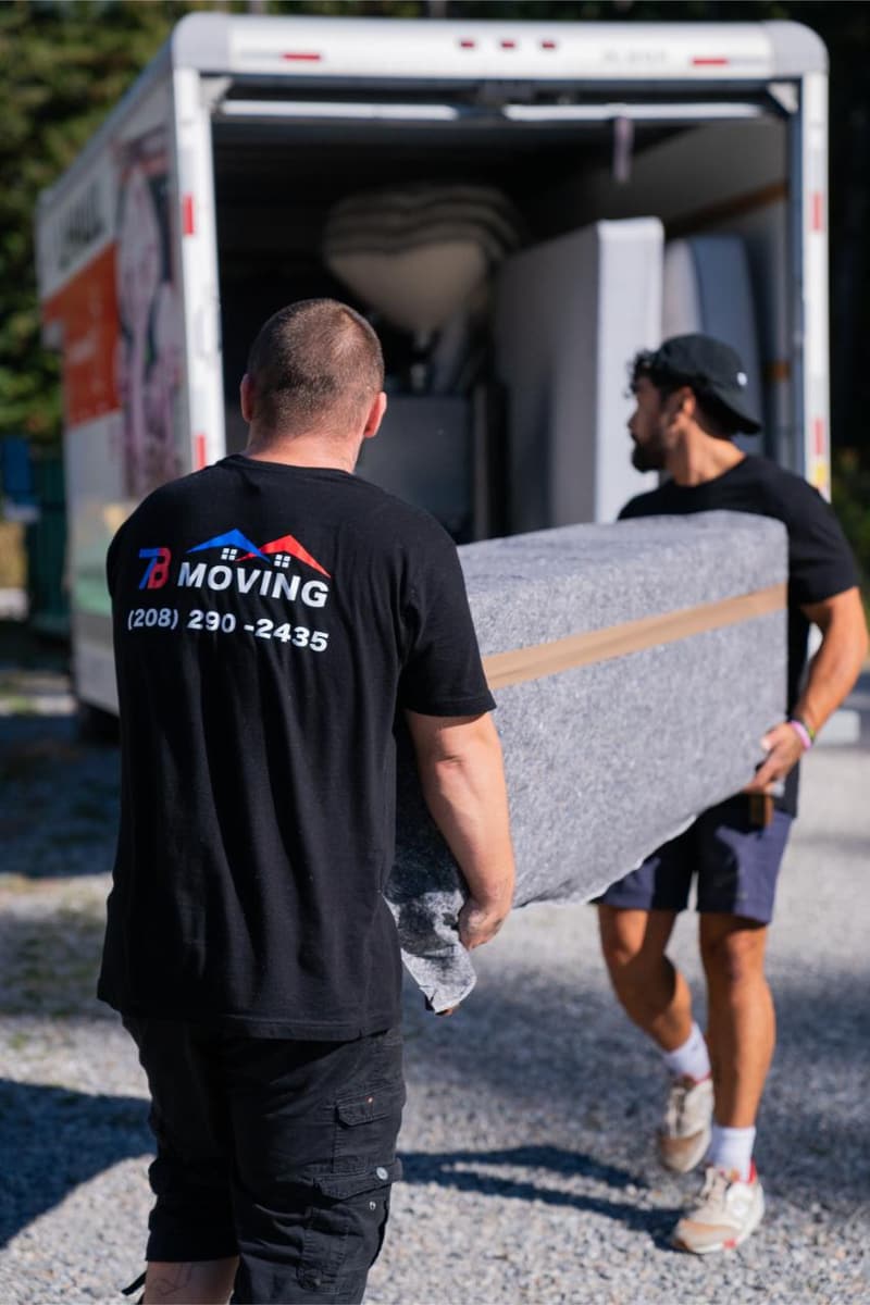 Two movers carry a pad-wrapped piece of furniture toward an open box truck