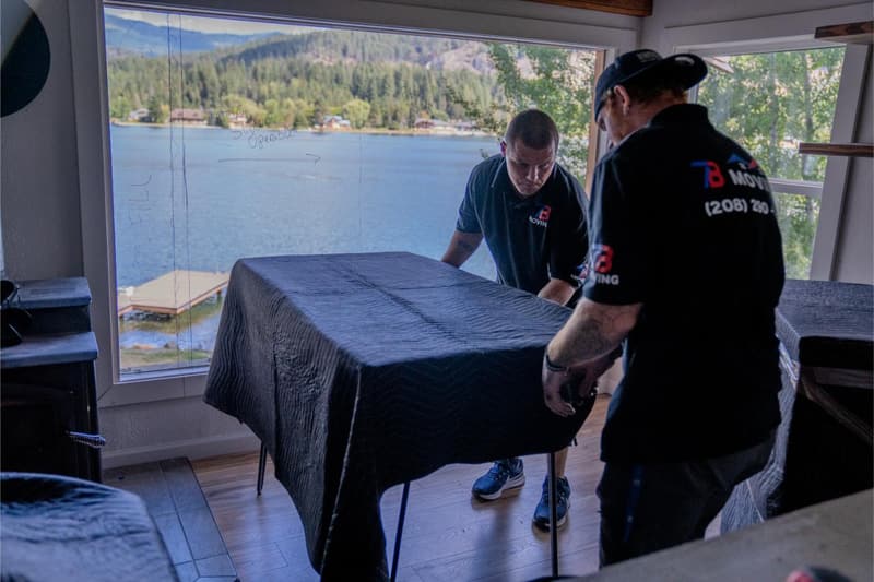 Two movers in 7B Moving shirts carry a blanket-covered table past a window overlooking water
