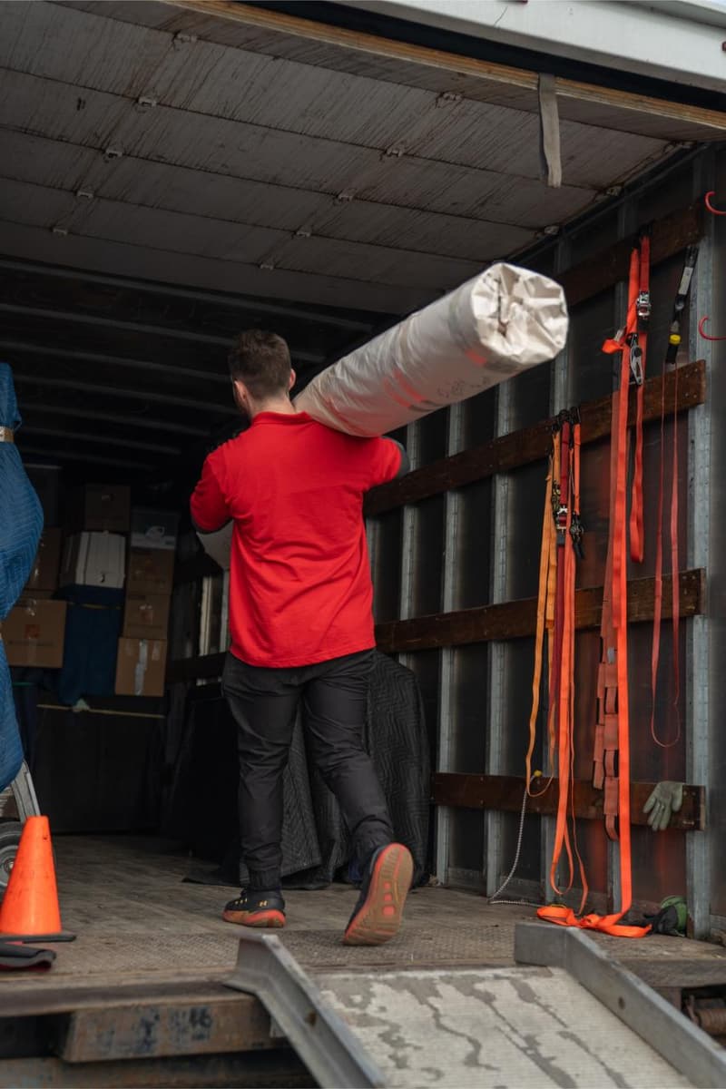 A mover carries a plastic-wrapped rolled rug up the ramp into a moving truck
