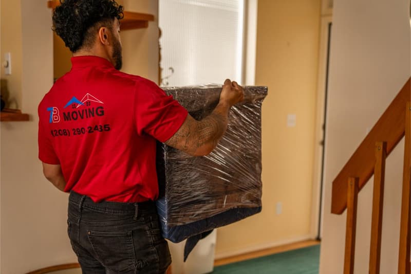 A mover in a red 7B Moving shirt carries a plastic-wrapped framed piece down a hallway