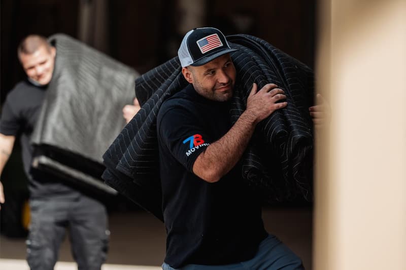 Two movers carry stacks of quilted moving blankets out of a garage