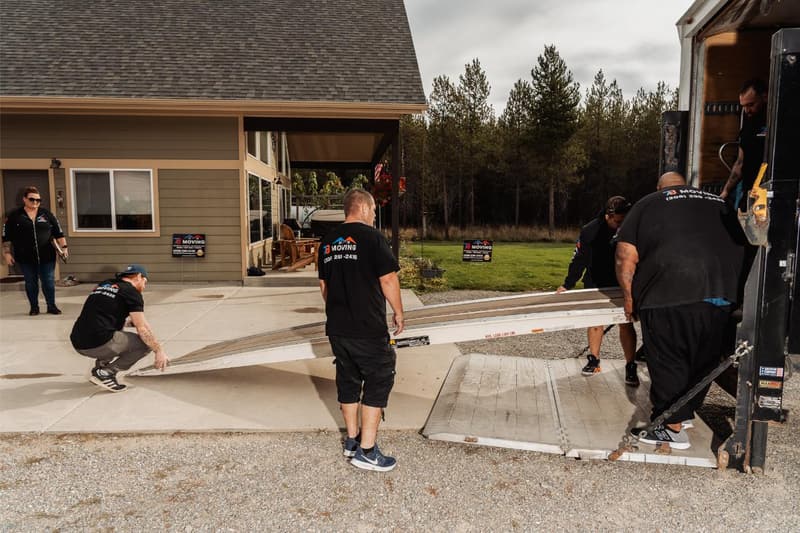 Movers in black 7B Moving shirts lower a loading ramp from the back of a box truck parked in a home's driveway