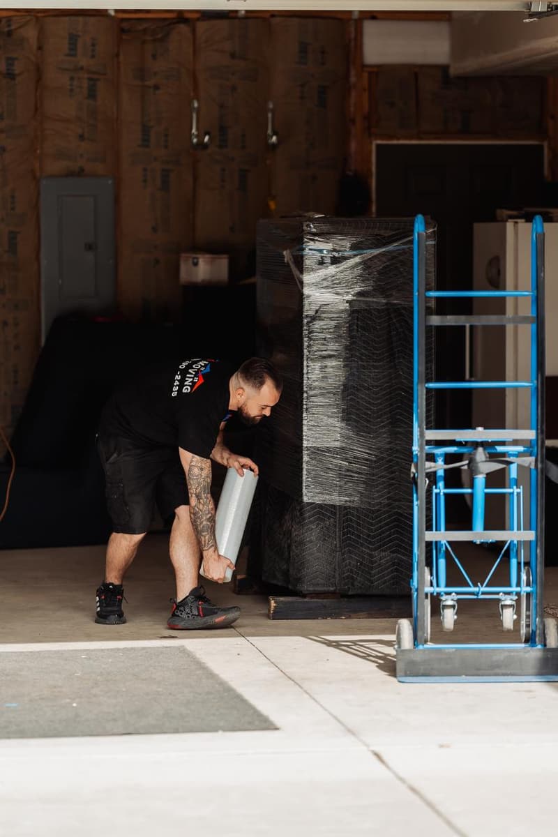 A mover kneels beside a wrapped tall item and a blue appliance dolly in an open garage doorway
