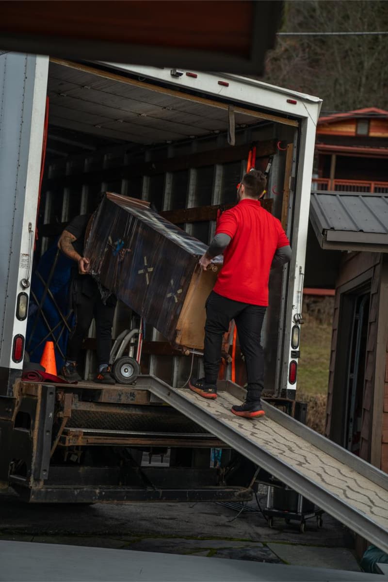 Movers roll wrapped furniture up a loading ramp into the back of a box truck