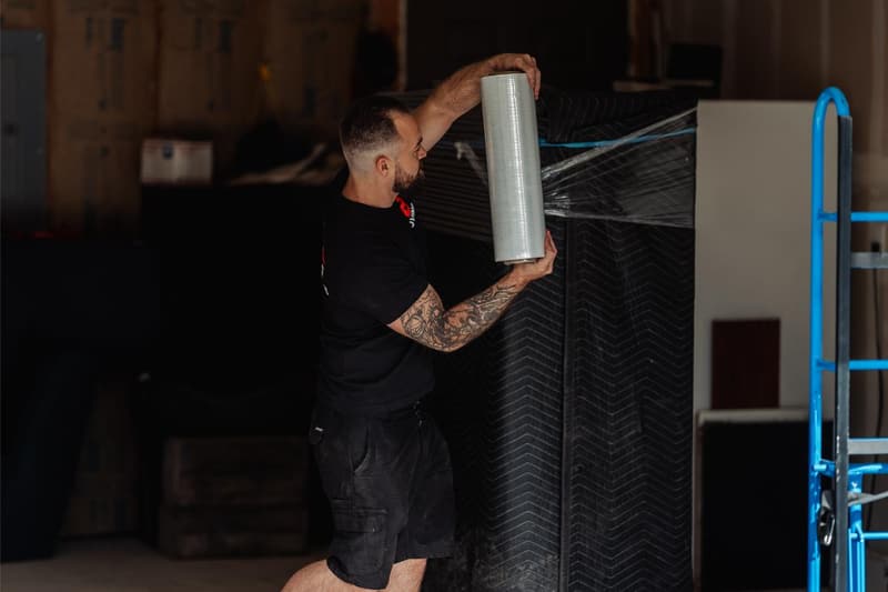 A mover stretch-wraps a blanket-covered cabinet beside a blue appliance dolly