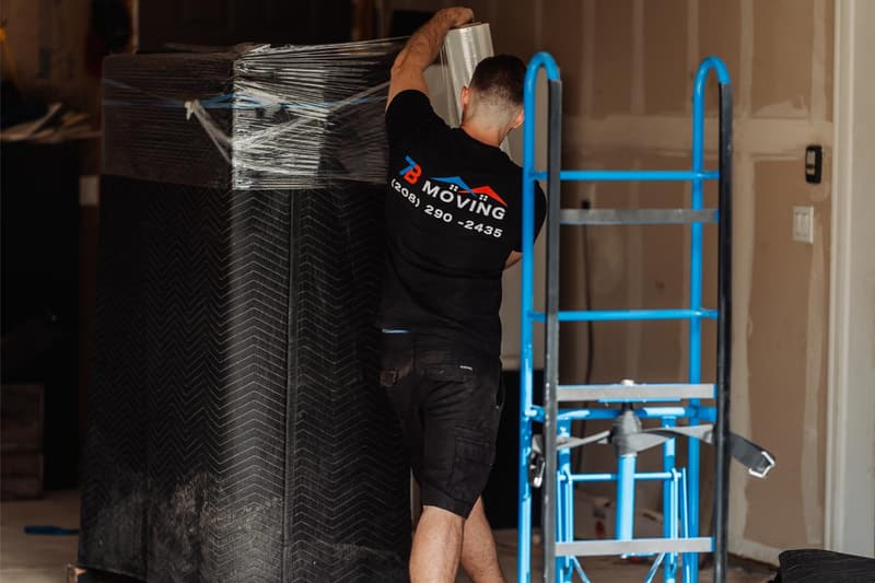 A 7B Moving crew member stretch-wraps a blanket-padded item next to a blue appliance dolly