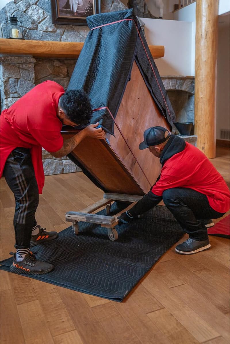 Two movers tip a wooden cabinet onto a moving pad to wrap it