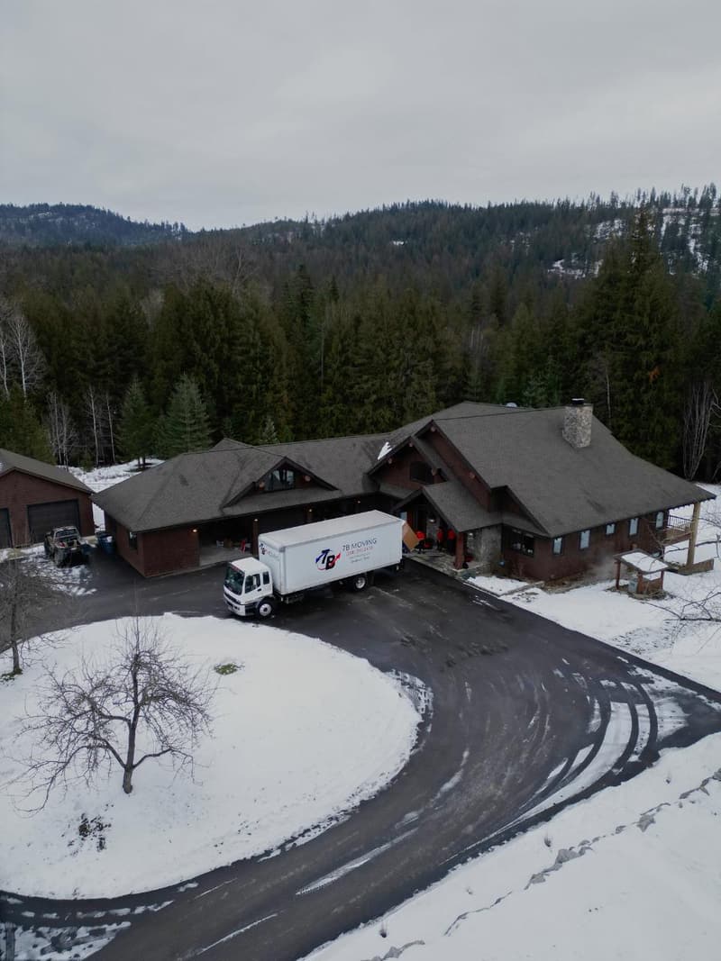 Aerial view of a moving truck parked on a curved driveway at a large house surrounded by snow and forest