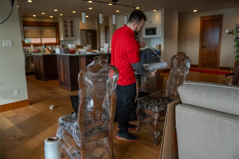 A mover in a red shirt wraps dining chairs in plastic inside an open kitchen