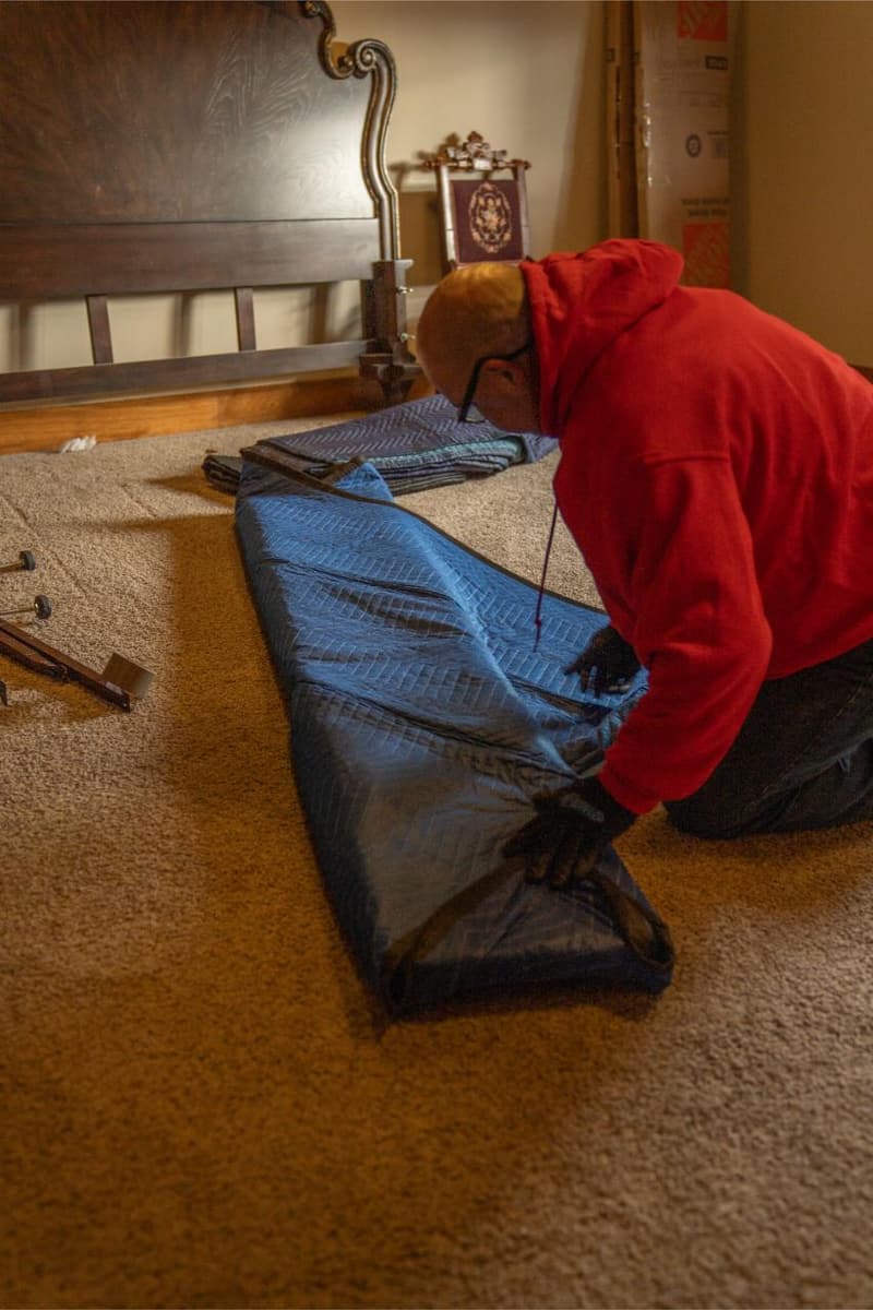 A mover in a red shirt kneels on carpet wrapping a long item in blue moving pads