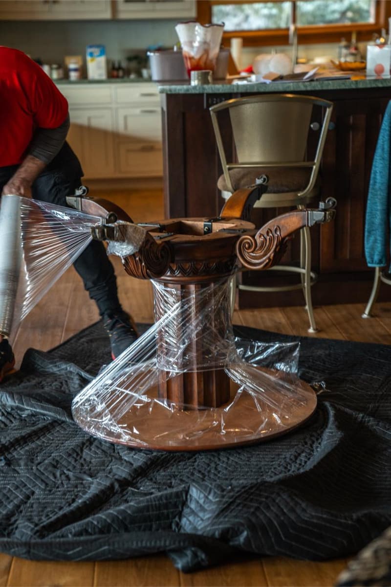 A carved wooden table base sits wrapped in clear plastic on moving blankets in a kitchen