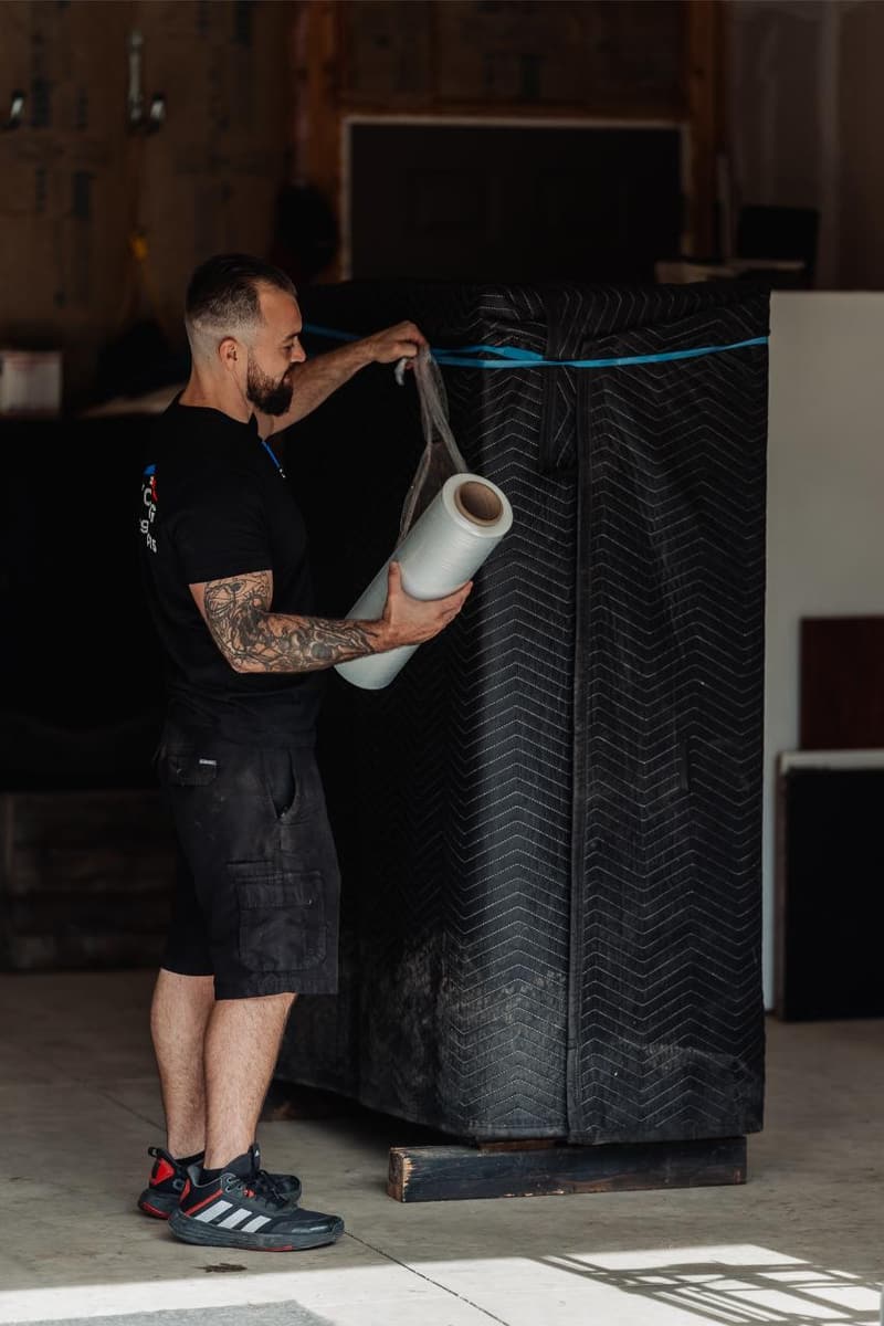 A mover stretch-wraps a tall blanket-covered item standing in a garage